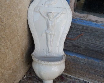 Puede incluir: Fuente de agua bendita de cerámica blanca montada en la pared con una representación de Jesucristo en la cruz. La fuente tiene un borde festoneado y un pequeño agujero en la parte superior para colgarla.