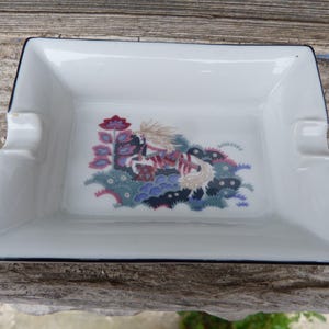 Könnte beinhalten: Weiße Aschenbecher aus Keramik mit schwarzem Rand. Der Aschenbecher zeigt ein farbenfrohes Blumenmuster mit einem Vogel in der Mitte.