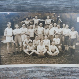 Peut inclure: Photographie vintage en noir et blanc d'un groupe de jeunes hommes, certains torse nu, posant en extérieur. Les hommes sont debout et assis sur l'herbe, avec un ballon au centre. La photo est légèrement délavée.