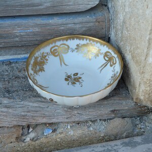 May include: A white porcelain bowl with a gold floral and ribbon design. The bowl is sitting on a wooden surface.