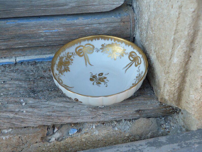 May include: A white porcelain bowl with a gold floral and ribbon design. The bowl is sitting on a wooden surface.