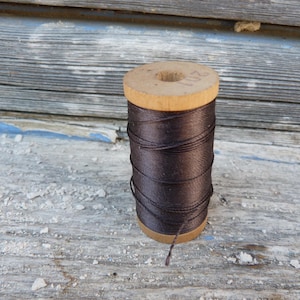May include: A wooden spool of dark brown thread with the number 201 stamped on the top edge. The thread is wound around the cylindrical wooden core, and a single loose strand hangs from the bottom of the spool against the wooden surface.