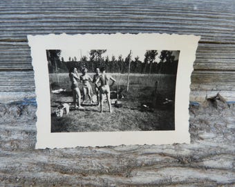 May include: Black and white vintage photograph of four people in swimwear standing in a grassy field. A small dog is near the group. Trees are in the background. The photograph has a decorative, scalloped edge.