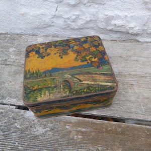 May include: Vintage square tin box with a landscape painting on the lid. The artwork depicts a bridge over a river, with trees and mountains in the background. The box has a yellow, orange, and green color scheme.