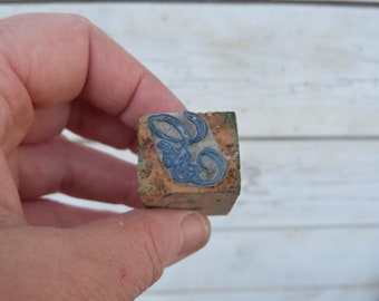 May include: A small wooden block with a blue and white design of a stylized floral pattern. The block is worn and has a rustic appearance.