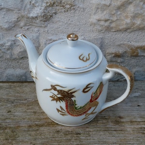 Può includere: Teiera in ceramica bianca con un design a drago dorato e dettagli dorati. La teiera ha un coperchio con un pomello dorato e un manico curvo. Il drago è raffigurato in oro, rosso e verde.