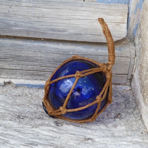 Puede incluir: Un flotador de vidrio azul oscuro, redondo, envuelto en una red tejida marrón, con un asa de cuerda a juego. El flotador descansa sobre una superficie gris desgastada, con un fondo de madera.