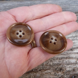 Puede incluir: Dos botones de cerámica marrón con un centro blanco y cuatro agujeros. Los botones son ligeramente brillantes.