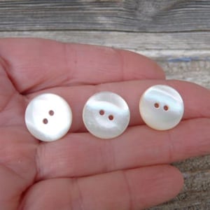 Op de afbeelding: Drie ronde, iriserende witte knopen met elk twee gaatjes. De knopen hebben een parelmoerglans die het licht reflecteert. Deze knopen zijn ideaal voor naaiprojecten en geven kleding een decoratieve uitstraling.