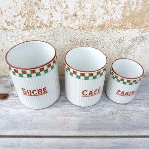 Puede incluir: Tres botes de cerámica blanca con bandas a cuadros verdes y naranjas y las palabras "SUCRE", "CAFE" y "FARINE" en letras marrones.