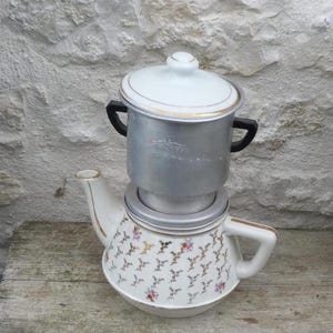 Puede incluir: Una cafetera vintage con una base de tetera de porcelana blanca adornada con un estampado floral y ribetes dorados. La parte superior es una cafetera de metal plateado con tapa. La cafetera está colocada contra una pared de piedra texturizada.