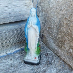 Op de afbeelding: Een wit gipsen beeld van de Maagd Maria, gekleed in een blauwe robe en met haar handen in gebed. Het beeld staat op een kleine basis met groene bladeren en rode bloemen.