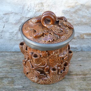 May include: Brown ceramic container with a lid, featuring a detailed relief of leaves and acorns. The container has a cylindrical shape with several small openings around the sides. The lid has a handle and a metal rim.
