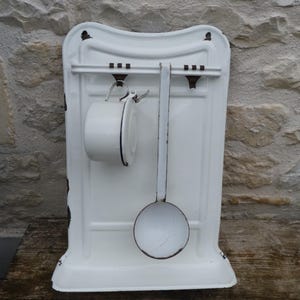 May include: White enamel kitchen wall rack with a hanging cup and ladle. The rack has a curved top and a horizontal bar for hanging utensils. The cup and ladle are also white with black trim, and the rack has some rust spots.