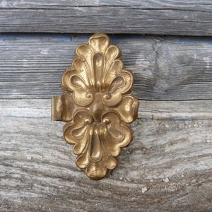 Puede incluir: Una pieza decorativa de metal dorado y ornamentado con un diseño floral. La pieza tiene una forma simétrica de hoja con detalles intrincados y un pequeño accesorio rectangular en un lado. Se encuentra sobre un fondo de madera gris desgastada.