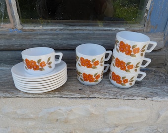 May include: Set of six white ceramic teacups with orange floral designs and matching saucers. The cups are stacked in pairs and the saucers are stacked in a pile.