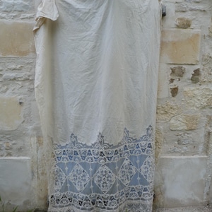 May include: A cream-colored, vintage-style textile with intricate lace detailing. The lower portion features a blue panel with a repeating diamond pattern, framed by delicate lace. The textile is hanging against a weathered stone wall.