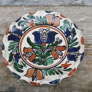 May include: A hand-painted ceramic plate with a scalloped edge. The plate features a central floral design in blue, orange, and green, surrounded by a brown border and additional floral motifs. The background is white, and the plate is set on a wooden surface.