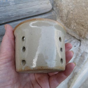 Op de afbeelding: Een cilindervormig keramieken vat met een glanzend, lichtbeige glazuur en een gespikkelde textuur. De buitenkant heeft twee verticale rijen van drie kleine, ronde gaten aan weerszijden van het lichaam, met een ongeglazuurde basisrand.