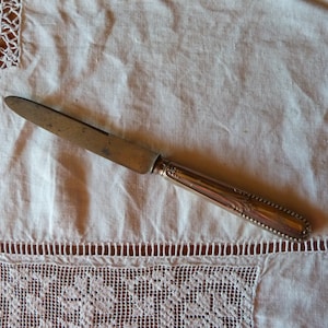 Puede incluir: Un cuchillo de mantequilla de plata con un mango adornado, sobre un mantel de lino blanco con ribete de encaje.