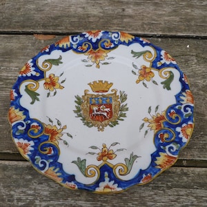 Könnte beinhalten: Ein weißer Keramikteller mit einem blauen und goldenen Blumendesign. In der Mitte des Tellers befindet sich ein Wappen mit einer Krone, einem Löwen und den Buchstaben "F.C."