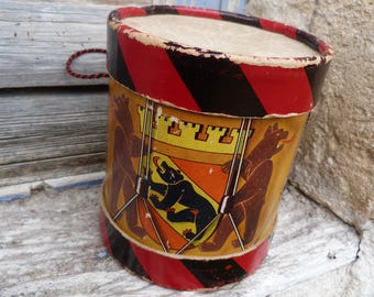 Puede incluir: Un tambor de juguete vintage de cartón con una banda roja y negra alrededor de la parte superior. El tambor está decorado con un diseño amarillo, naranja y rojo que presenta tres osos y un castillo.