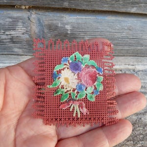 May include: A square, red, perforated plastic canvas piece featuring a central, colorful floral bouquet illustration. The bouquet includes a white daisy, a pink rose, and blue flowers with green leaves, all tied together with a small red ribbon.