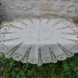 Può includere: Tovaglia rotonda color crema con bordi in pizzo intricati. Il centro presenta dettagli floreali ricamati. La tovaglia è esposta su una superficie di legno invecchiato, mostrando il suo design delicato e l'estetica vintage.