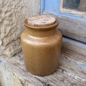 Puede incluir: Un tarro de cerámica marrón con tapa de corcho. El tarro tiene un esmalte marrón moteado y está sentado sobre una superficie de madera.