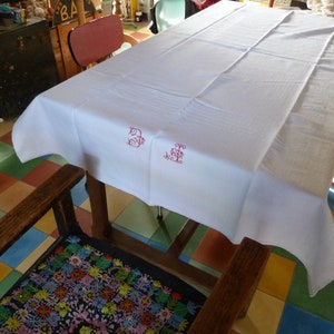 May include: White tablecloth with embroidered initials on a table with a colorful checkered floor. The tablecloth is white with a subtle texture and has two embroidered initials in pink thread. The floor is a checkered pattern of yellow, green, blue, and orange tiles.