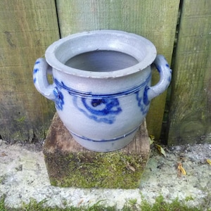 Peut inclure: Un pot en grès gris clair doté d'un large rebord, de deux anses latérales et d'un motif floral bleu cobalt peint à la main entourant le corps. Le récipient présente une forme cylindrique avec une base légèrement effilée et une finition émaillée.