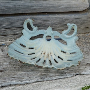Può includere: Un portasapone in ghisa blu chiaro con un design a conchiglia. Il piatto ha un bordo smerlato e volute decorative. La vernice è usurata, conferendogli un aspetto vintage. Il piatto misura circa 15 cm di larghezza.