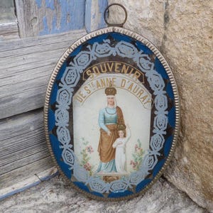 Puede incluir: Un recuerdo ovalado con un marco azul y plateado. La imagen interior representa dos figuras, una con un vestido azul y una túnica marrón, la otra de blanco, con el texto "SOUVENIR DE STE ANNE D'AURAY" en la parte superior.