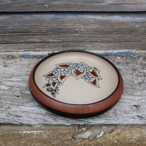 May include: A brown ceramic bowl with a black rim and a white floral design. The design features brown, white, and blue leaves.
