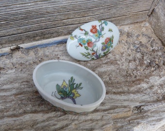 Może przedstawiać: Białe pudełko na drobiazgi z porcelany z kwiatowym wzorem na pokrywce i wzorem motyla na spodzie. Pudełko ma owalny kształt i błyszczące wykończenie.