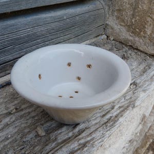 May include: A white ceramic bowl with several small holes, likely for drainage. The bowl has a rounded shape and sits on a weathered wooden surface. The background includes a section of weathered wood and stone.