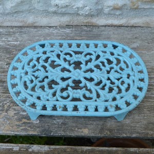 Puede incluir: Un salvamanteles de hierro fundido azul claro, de forma ovalada y con un diseño floral calado y ornamentado. El salvamanteles tiene tres pequeños pies y está sobre una superficie de madera envejecida.