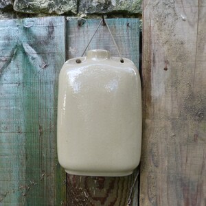 Può includere: Una brocca in ceramica beige con un manico e una corda attaccata alla parte superiore, appesa a una recinzione di legno dipinta di blu e verde.