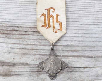 May include: A silver religious medal with a cross design and an image of Jesus. The medal is attached to a white ribbon with the letters "IHS" embroidered in gold.