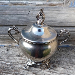 Op de afbeelding: Een zilverkleurige suikerpot met een deksel en twee handvatten. De kom staat op een decoratieve voet met sierlijke details. Het deksel heeft een gebogen handvat. De kom is reflecterend.