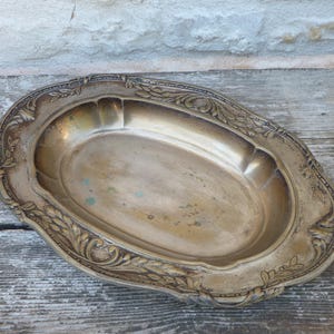 Op de afbeelding: Een ovaalvormig dienblad van messing met een diepe centrale kom en een brede, decoratieve rand. De rand is versierd met ingewikkelde reliëfpatronen van bladeren en krullen, met een verweerde, verouderde patina en subtiele groene oxidatievlekken.