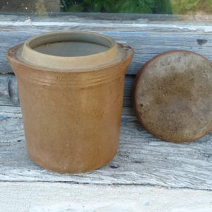 Op de afbeelding: Een bruine keramische pot met deksel, waarschijnlijk voor voedselopslag. De pot is cilindrisch met een licht uitlopende rand en twee kleine handvatten. De deksel is rond en plat. De pot en deksel hebben een rustieke, verouderde uitstraling.