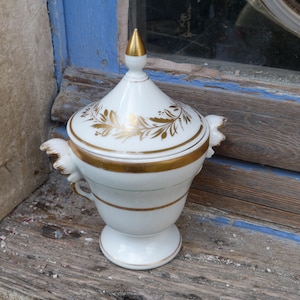 May include: White porcelain sugar bowl with a gold lid and gold floral design. The bowl has two handles and sits on a pedestal base.