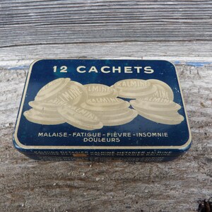 Peut inclure: Une boîte en métal bleue vintage avec l'inscription "12 CACHETS" en blanc. La boîte présente une illustration de plusieurs pilules blanches et du texte en français, dont "MALAISE-FATIGUE-FIÈVRE-INSOMNIE DOULEURS".