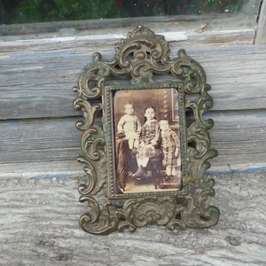 May include: Ornate antique brass picture frame with a sepia-toned photograph of three children. The frame has an elaborate, scrolling design and a weathered, aged appearance. The photograph is rectangular and centered within the frame.