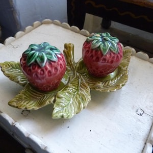 Può includere: Un set di saliera e pepiera a forma di fragola in ceramica. I shaker sono rossi con cime verdi, appoggiati su una base verde a forma di foglia. Il set è posto su un tavolo bianco con bordo smerlato.