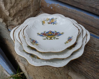 May include: A set of three white ceramic plates with scalloped edges and gold trim. The plates feature a floral design with blue, yellow, and red flowers.