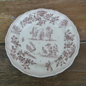 Könnte beinhalten: Ein weißer Keramikteller mit braunem Blumendesign und einer Szene mit Figuren und Vögeln. Der Teller hat einen gewellten Rand.