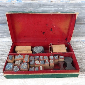 Könnte beinhalten: Eine offene, rote Vintage-Box mit grünem Rand, gefüllt mit Holzstempeln. Die Stempel umfassen Buchstaben, Zahlen und dekorative Designs. Einige Stempel haben blaue Tinte. Die Box steht auf einer verwitterten Holzoberfläche.
