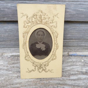 May include: Antique sepia-toned portrait of a person in a dark dress and white bonnet, set within an oval frame. The frame is surrounded by ornate, cream-colored embellishments on a rectangular card.
